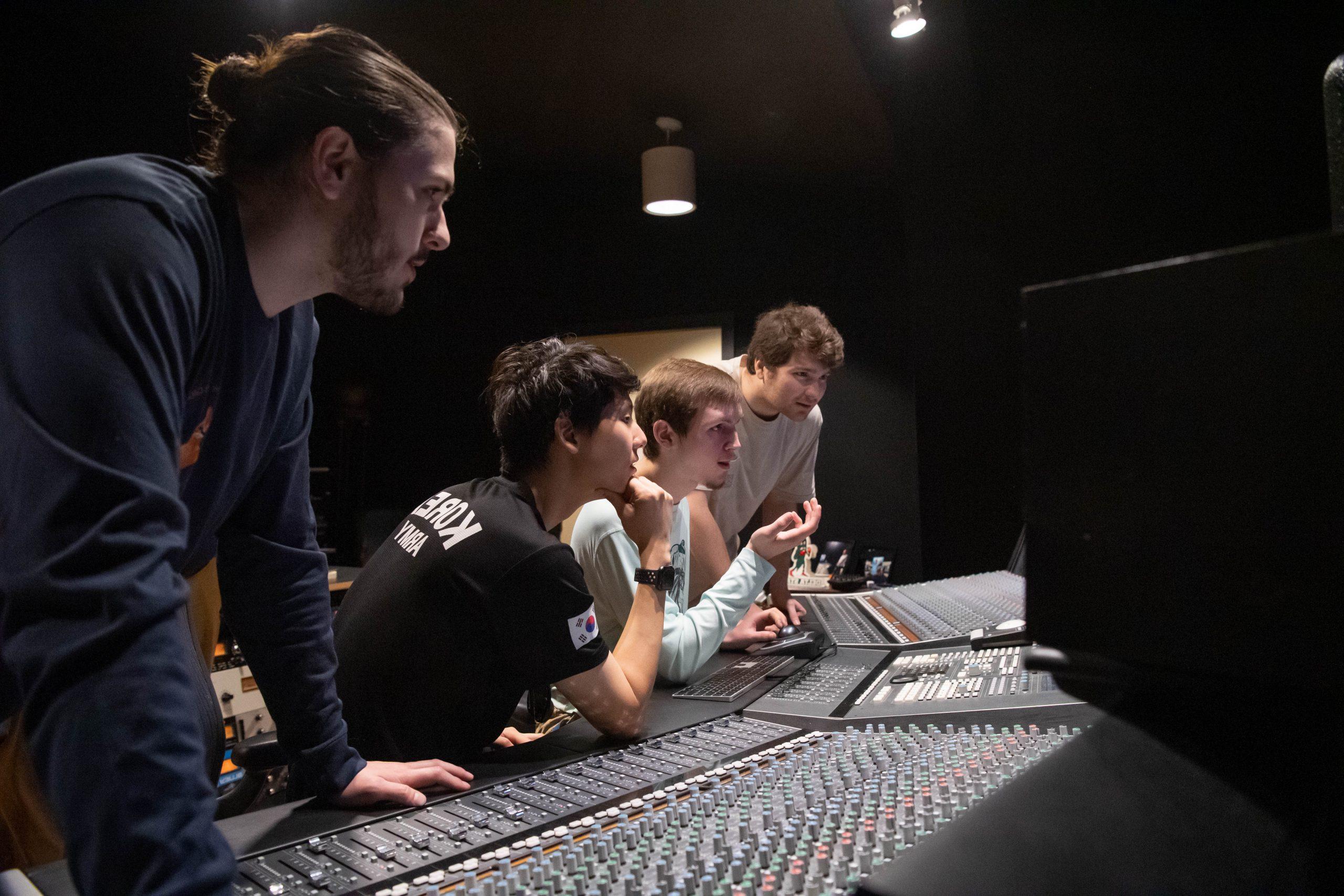 四个学生站着或坐在录音室的控制台前，在做一个项目.