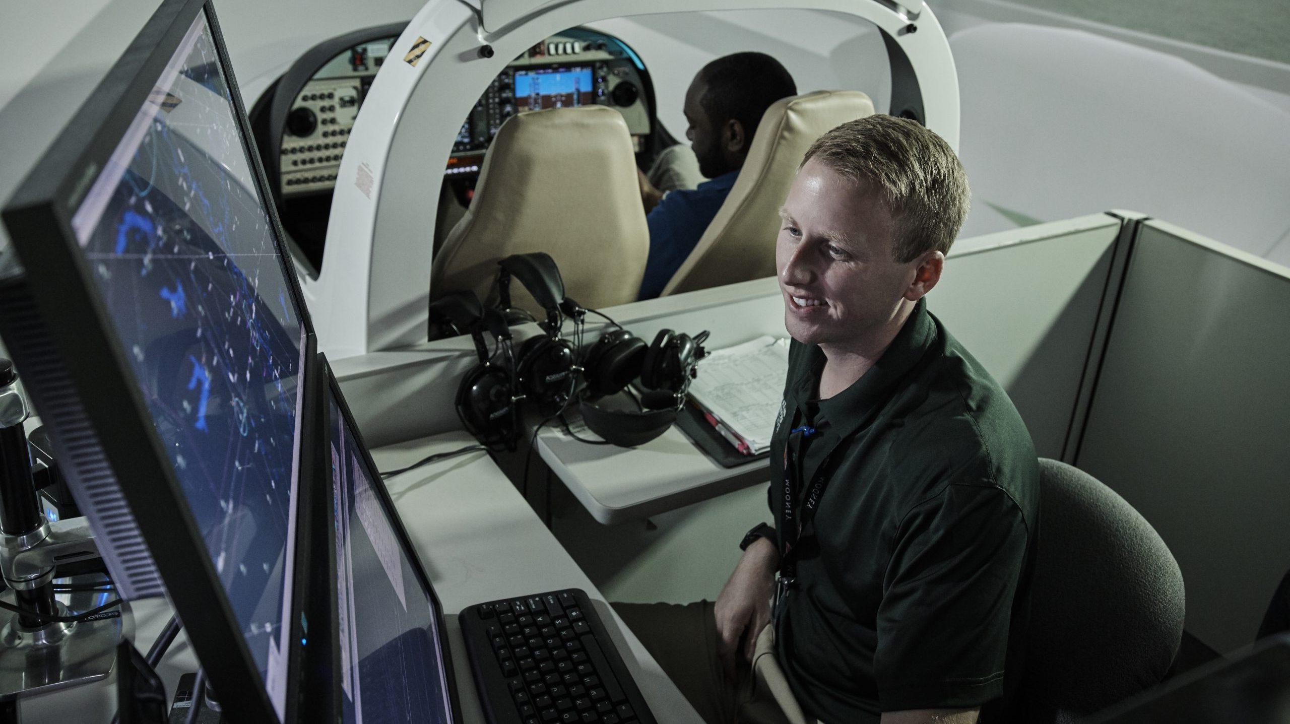 看着电脑屏幕的学生, 监察空中交通, 而另一个学生在他身后的飞行模拟器里.