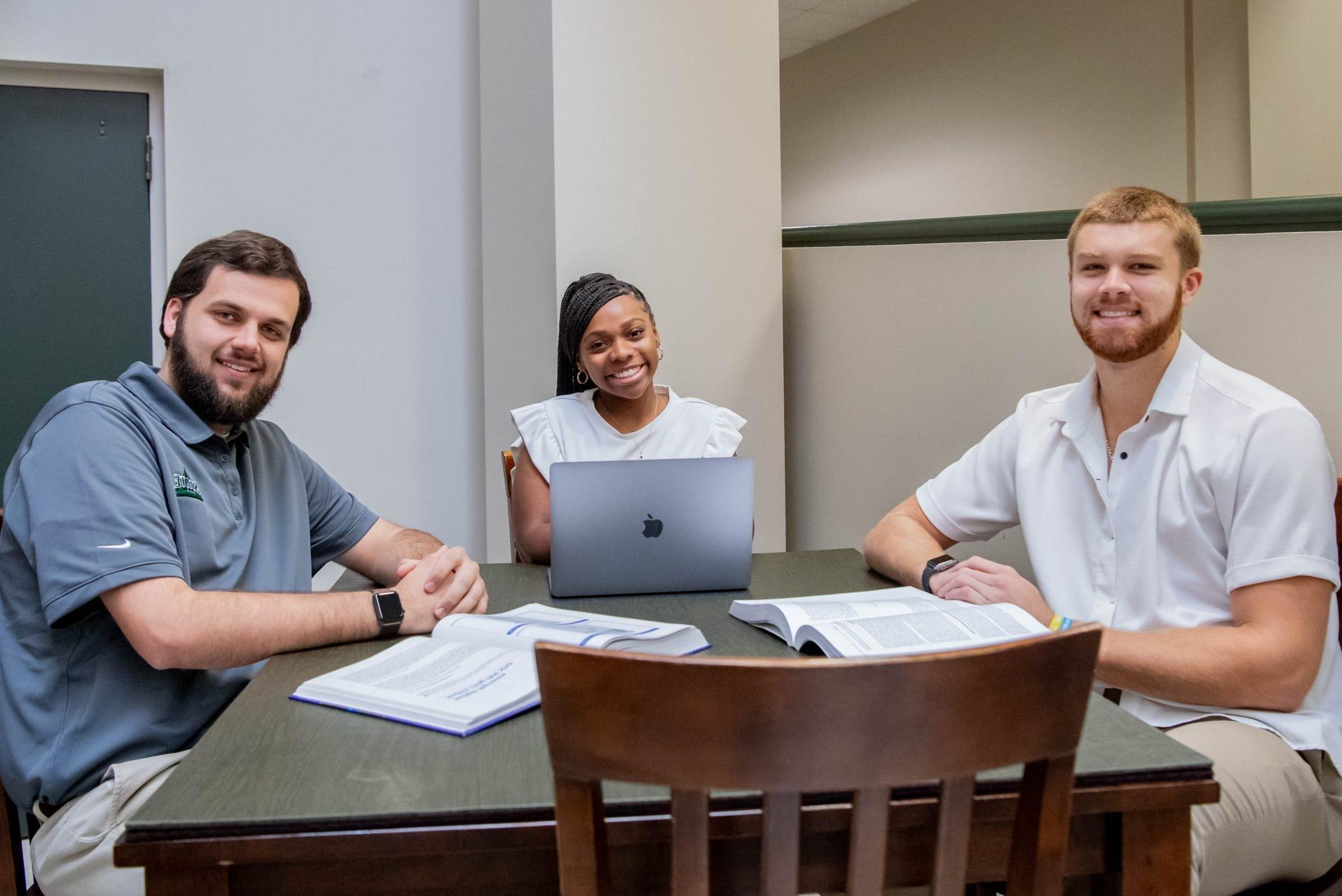 Three students working on assignment together in the College of Business and Aviation lobby, with a laptop and two textbooks.